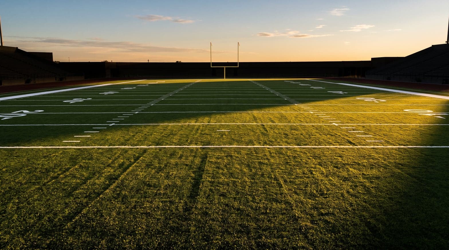 Campo de futebol americano vazio ao fim da tarde, com linhas de jarda e trave ao fundo